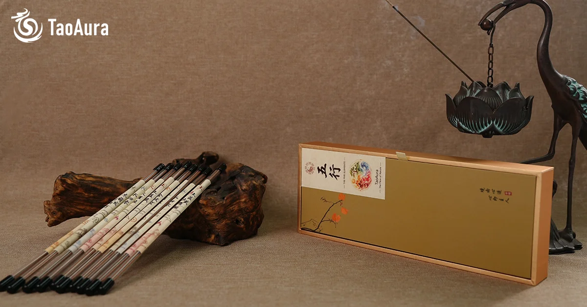 Five elements incense stick blend — color-coded Wu Xing sticks in green red beige white and dark on gnarled wood, wooden box with 五行 diagram plum blossom and calligraphy, bronze crane lotus censer with burning smoke — TaoAura Wood Fire Earth Metal Water feng shui ritual